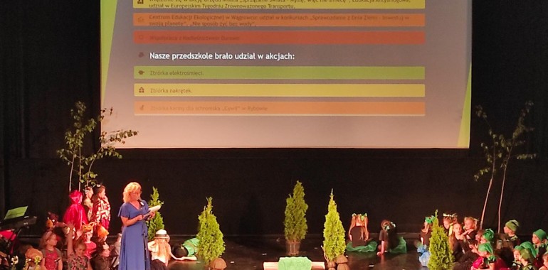 Zdjęcie z wydarzenia: Podsumowanie projektu „Przedszkole jako ośrodek zrównoważonego rozwoju” - przedstawia Panią Dyrektor z dziećmi na scenie